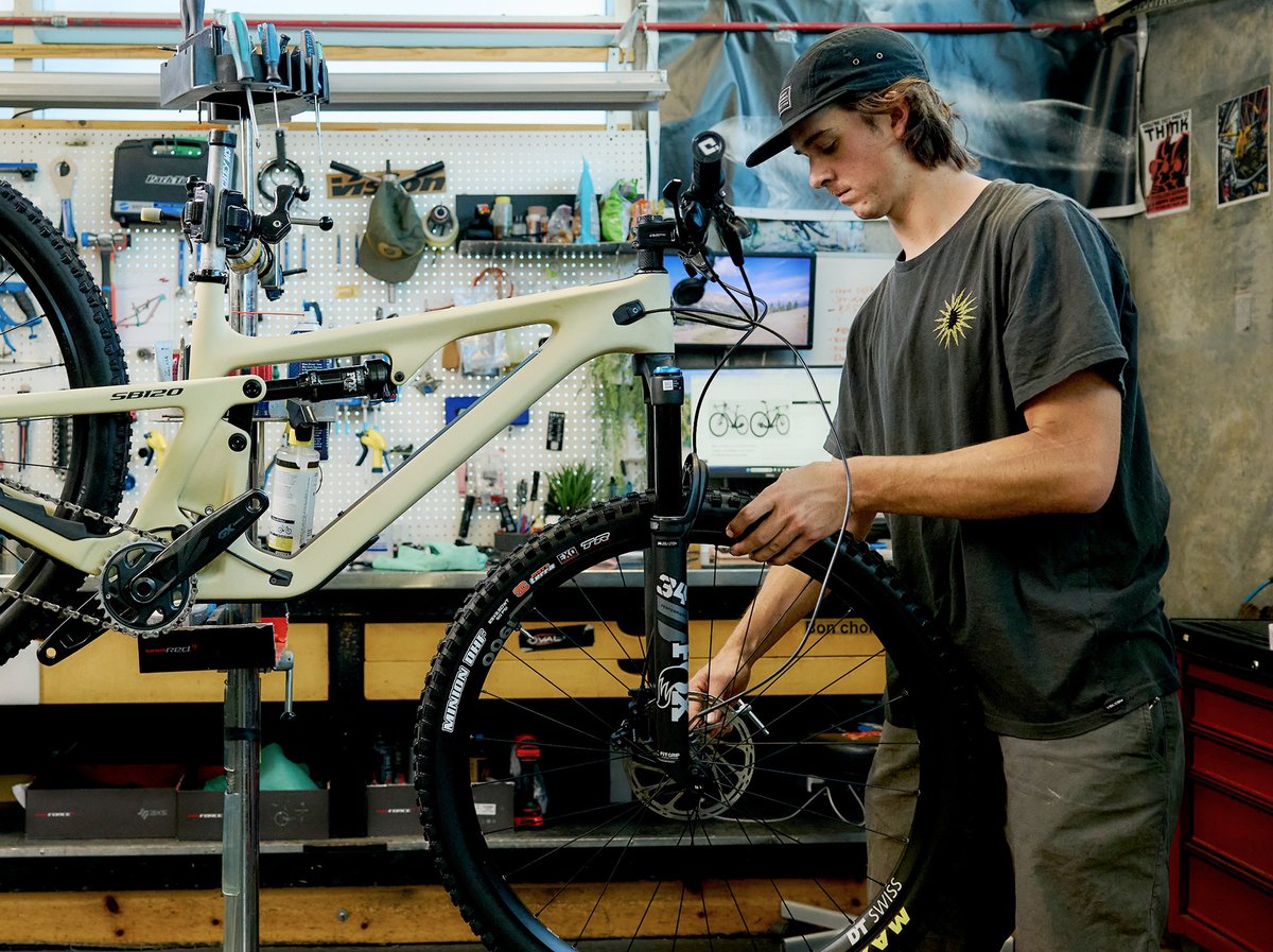 A man in a bike shop stands over a bike frame. He’s looking at the camera with a face that says, “I can build any bike, try me.” 