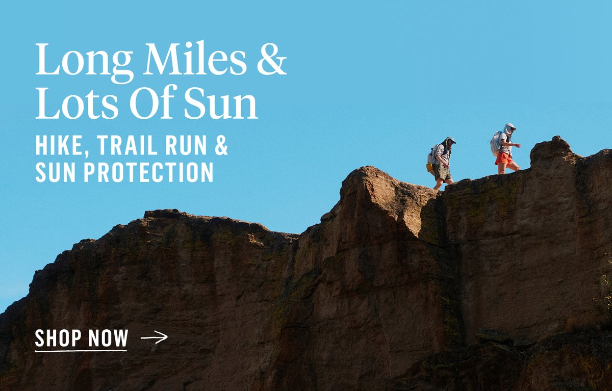 Two hikers traversing a sun-exposed sandstone ridgeline with loaded packs against a clear blue sky.