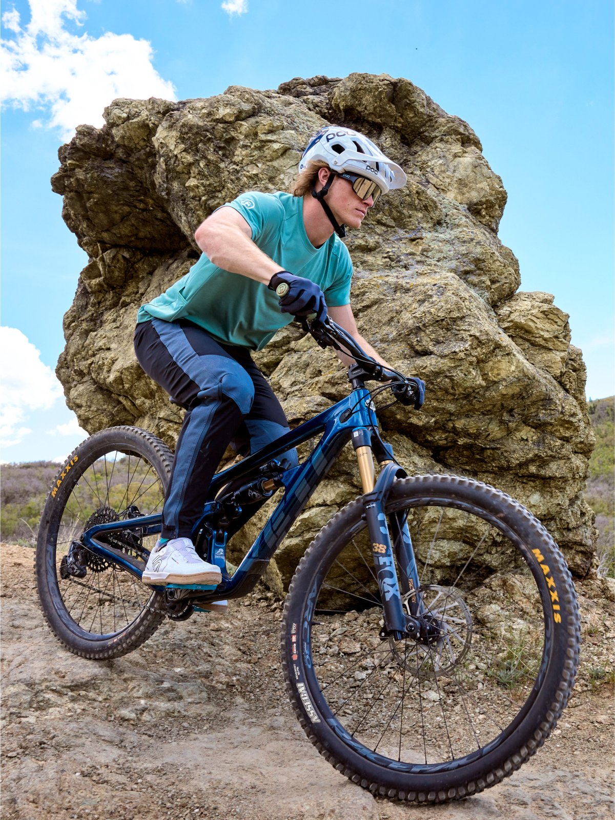 A mountain biker wearing a white helmet, teal T-shirt, black gloves, and blue athletic pants rides a blue mountain bike on a rocky trail with a large rock formation and blue sky in the background.