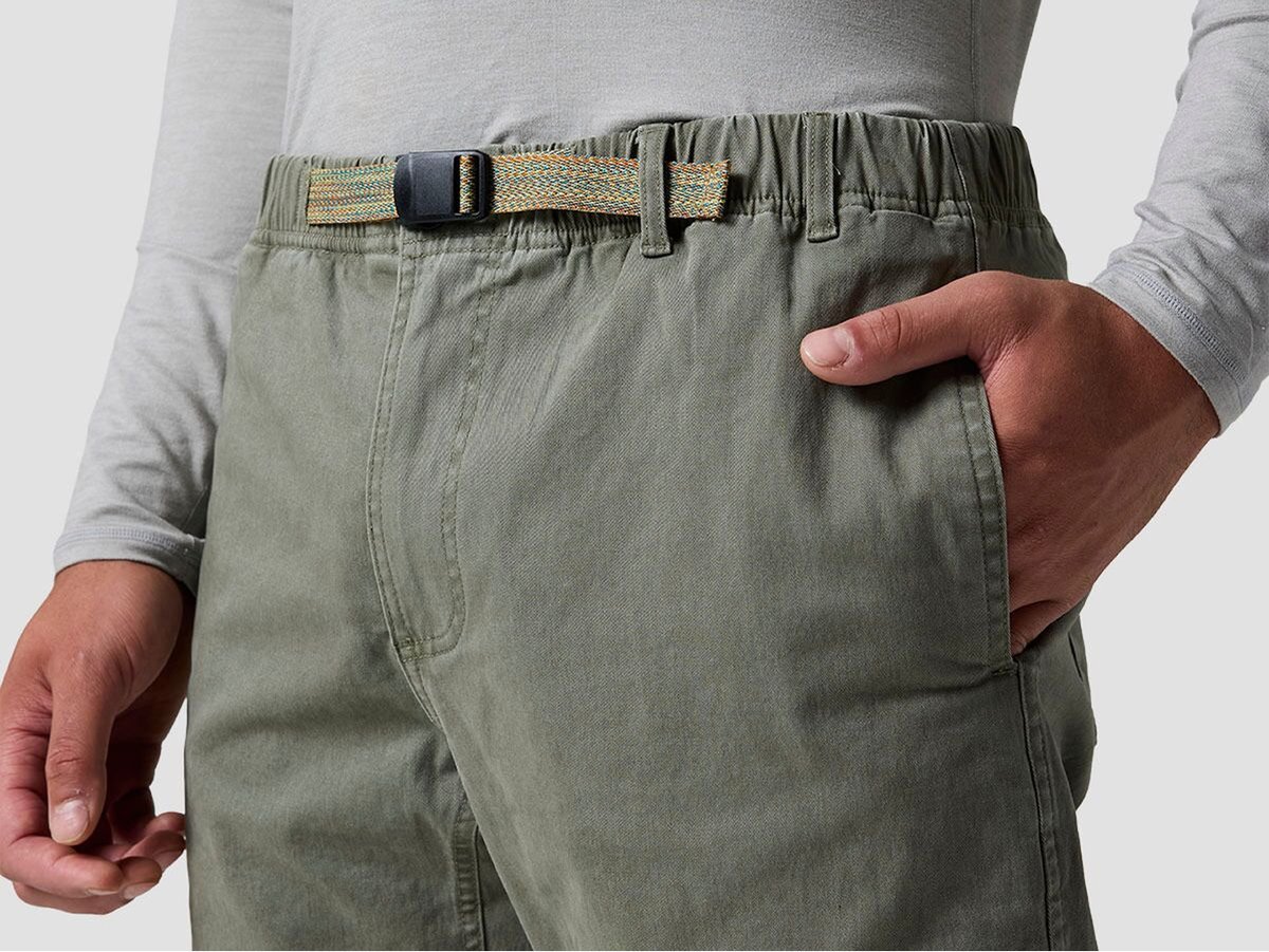 Close-up of a person wearing sage green pants with a woven belt, hand resting in the front pocket.