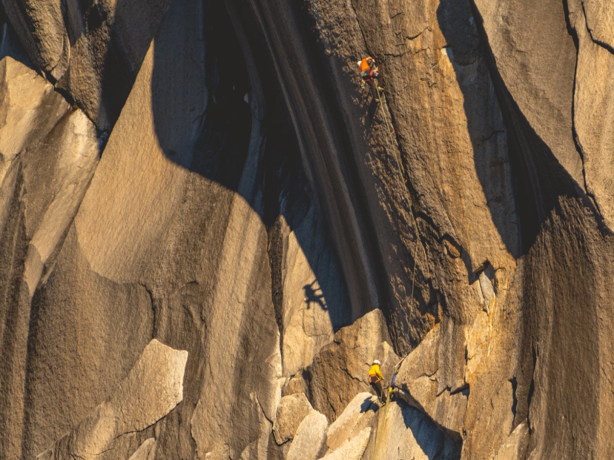 Two alpinists wearing tech hoodies, shorts, hiking boots, and running vests scale a bouldery ridge.