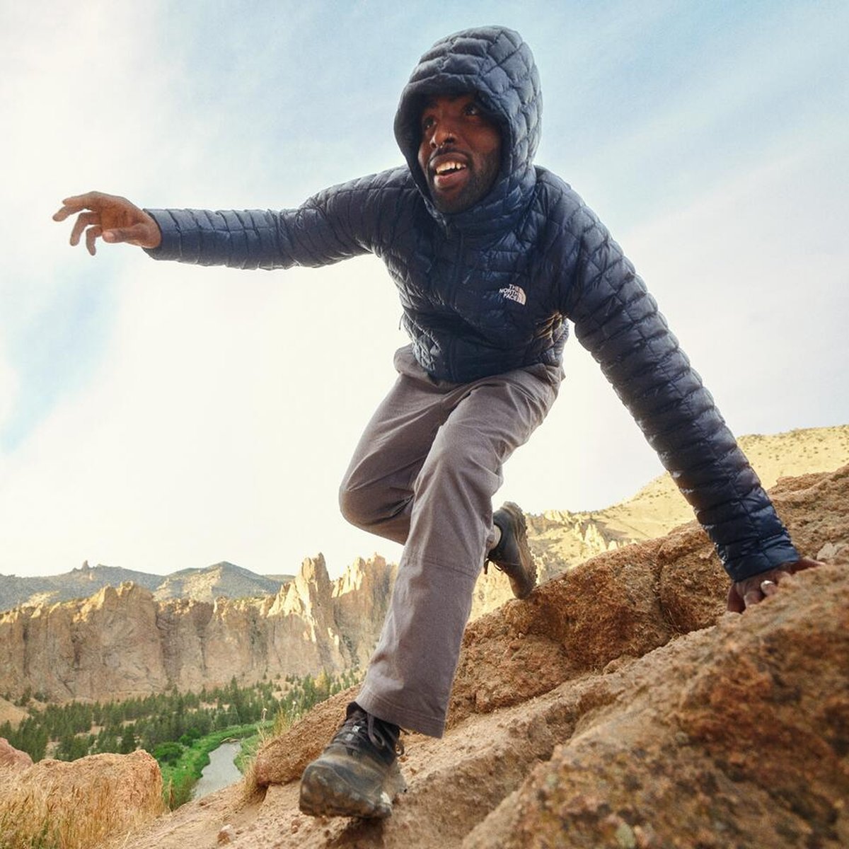 A man in a hooded puffer jacket climbs up a rocky slope with a canyon in the background.