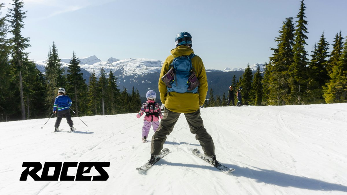 An adult skier in a yellow jacket instructs a young child in pink skis on a snowy mountain slope surrounded by evergreen forests and snow-capped peaks.
