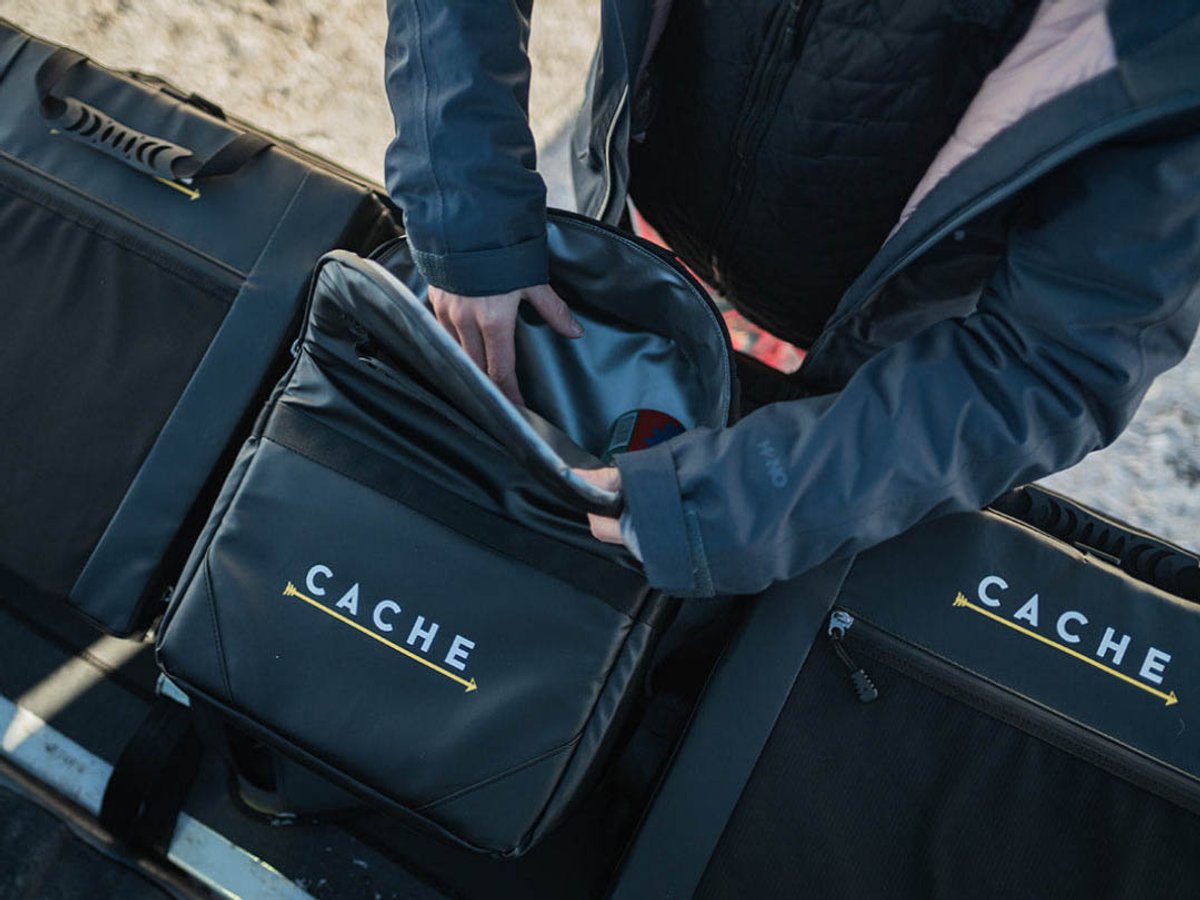 Person loading a black CACHE gear bag into a larger CACHE storage case mounted in a truck bed.