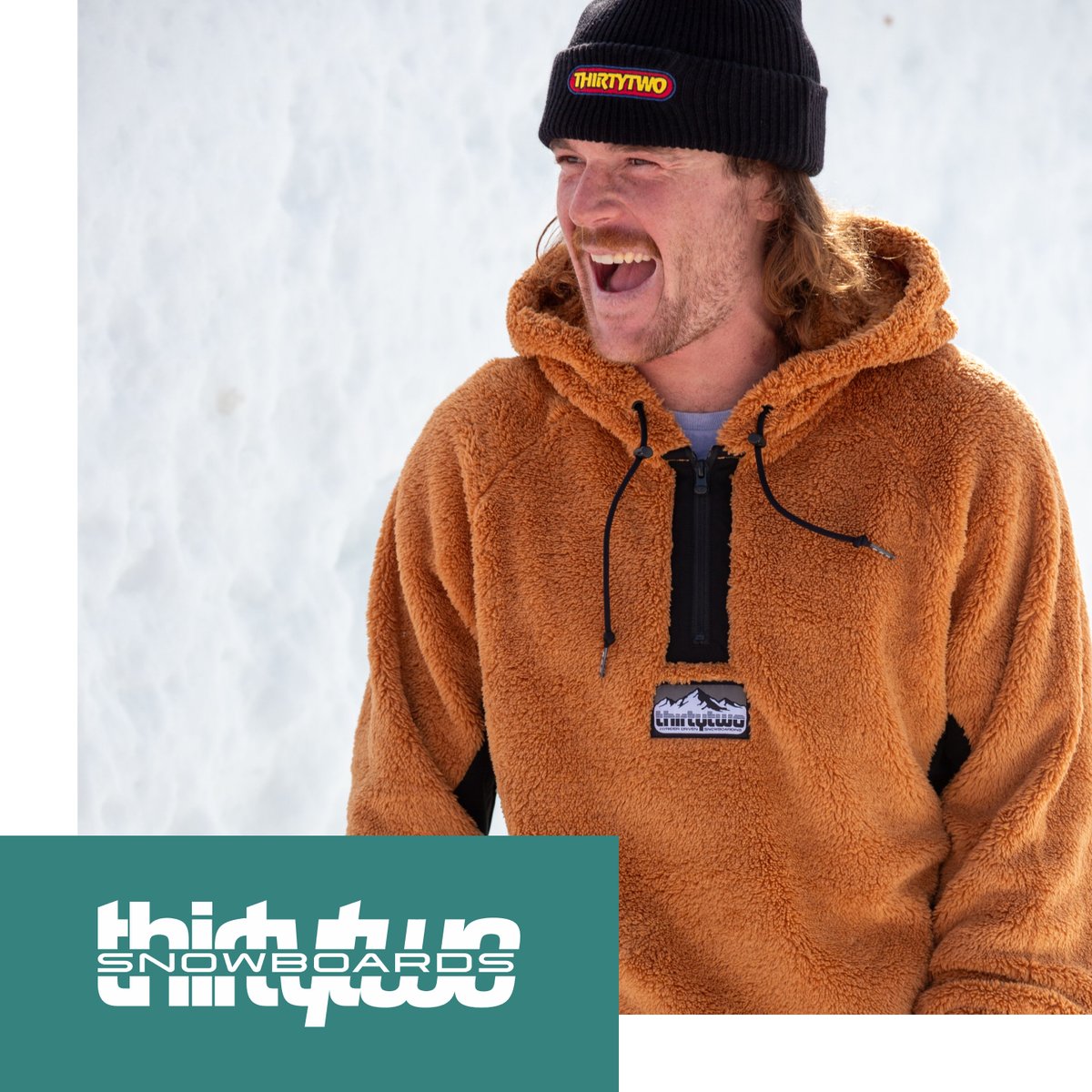 Shop thirtytwo Snowboards — man wearing black thirtytwo beanie and tan fleece hoodie, smiling in snowy setting
