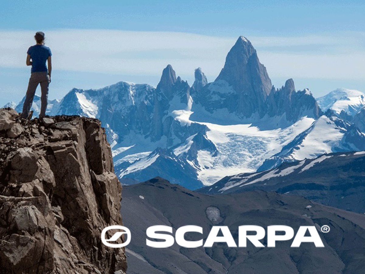 Scenic mountain landscape with a person standing on a rocky cliff overlooking snow-covered peaks and a dramatic alpine spire, with the SCARPA logo displayed across the bottom.