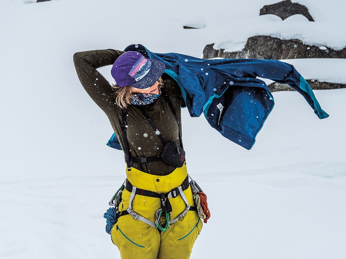 Climber pulling on a blue shell jacket mid-snowstorm, wearing a climbing harness loaded with carabiners and yellow mountaineering pants.