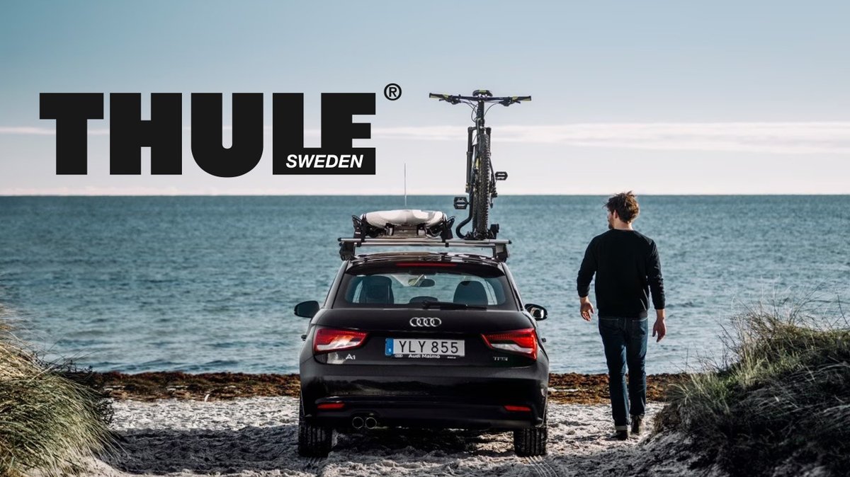 A man stands on a beach next to a black car equipped with a Thule roof rack carrying a bicycle and gear.