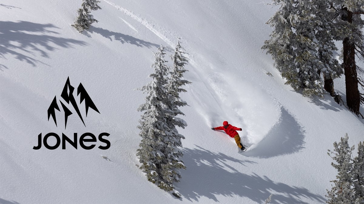 Snowboarder in red jacket carving through deep powder snow on a steep mountain slope lined with snow-covered evergreen trees, showcasing Jones snowboard gear performance.