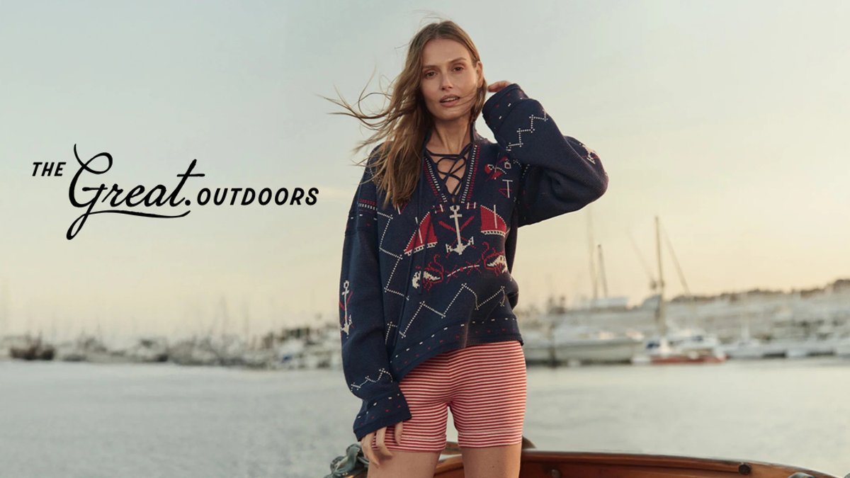 Woman wearing navy nautical-patterned hoodie and red striped shorts standing on a boat with sailboats in the background for The Great Outdoors brand.