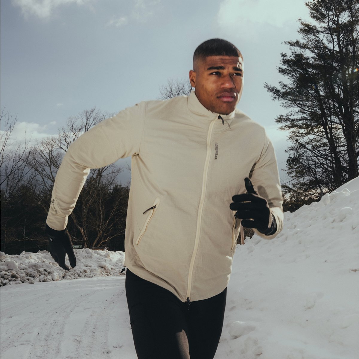A person wearing a light jacket and black gloves is running on a snowy path with trees and cloudy sky in the background.