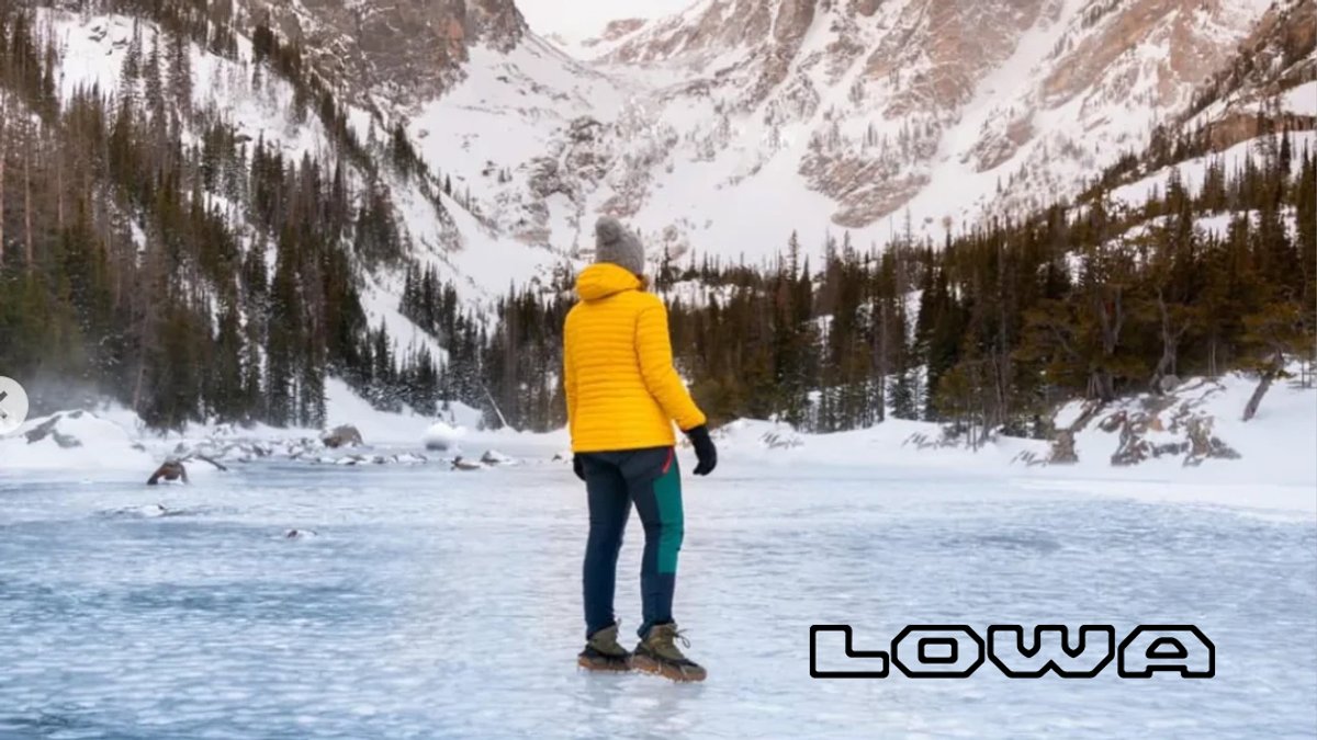 A runner in a bright yellow jacket trails along a frozen alpine lake surrounded by snow-covered mountains and evergreen forests.