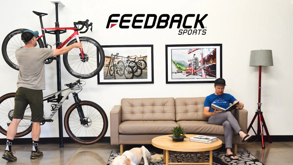 Modern bike storage solution displayed in Feedback Sports showroom with cyclist mounting red road bike on wall-mounted rack while person reads on couch surrounded by cycling photography and gear.