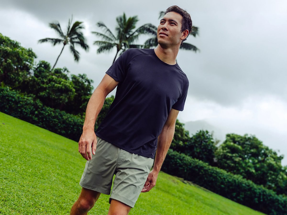 Man wearing the Backcountry Wasatch Merino T-Shirt in navy with khaki shorts, standing on a grassy hillside with palm trees and a stormy tropical sky in the background.