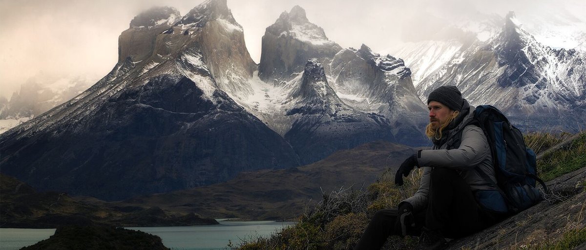 Person sitting on grassy slope wearing Coalatree Camper Jacket in front of majestic snowy mountain peaks.