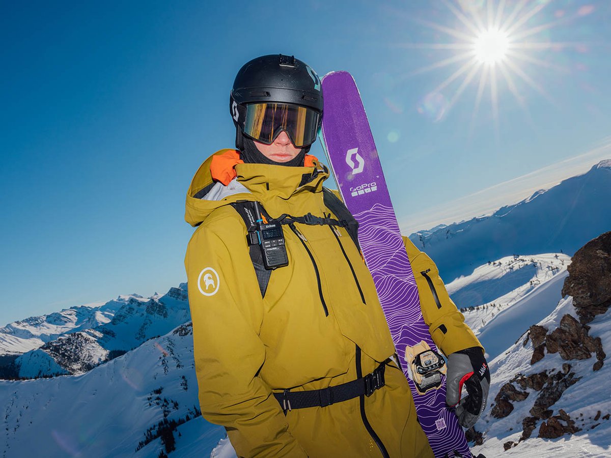 Skier holding skis wearing backcountry jacket and backpack in snowy alpine space with sun shining in the background.