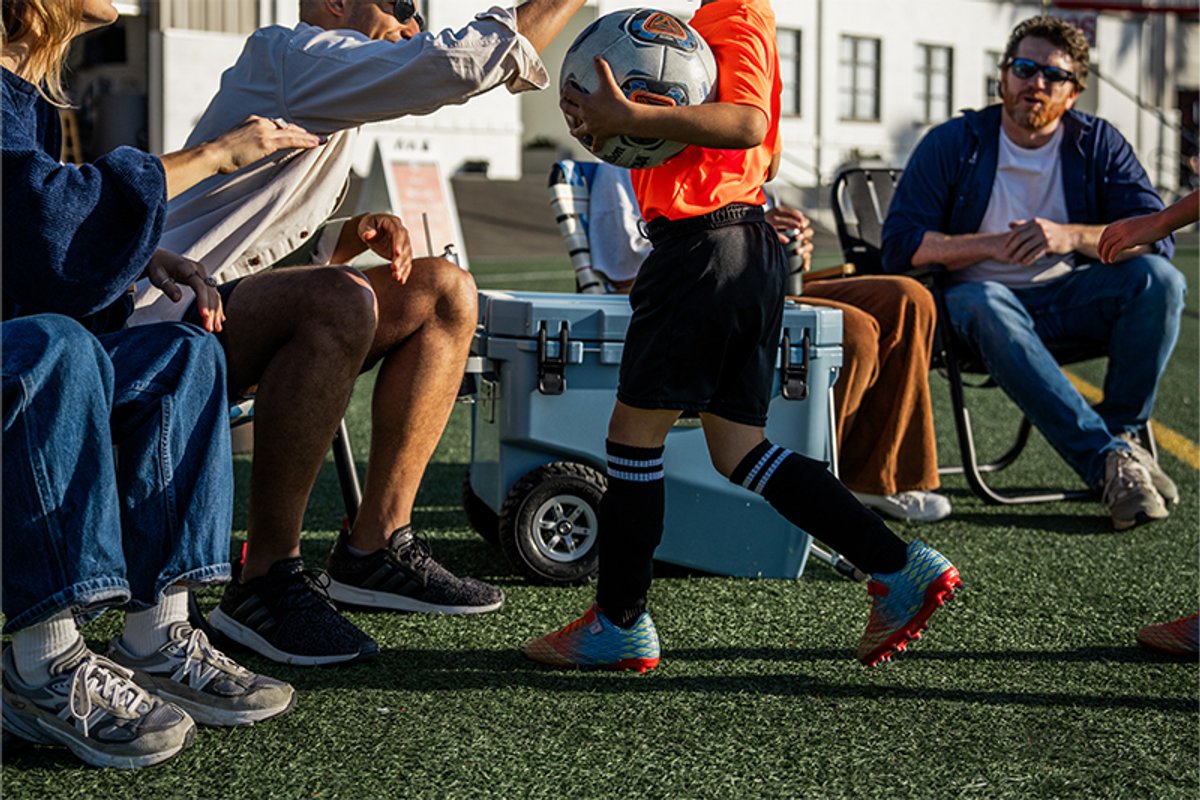 A family sits on a soccer pitch in camp chairs. A child runs over to them, and a man gives him a high five. The RovR RollR cooler sits next to the family.