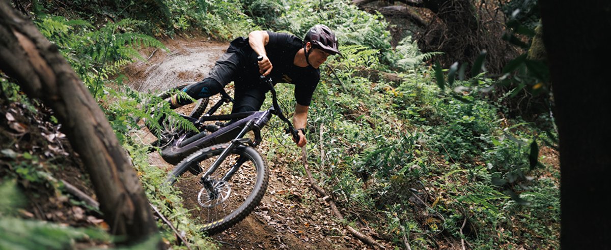 Mountain biker charging steep overgrown forest descent on full-suspension MTB surrounded by roots and green vegetation