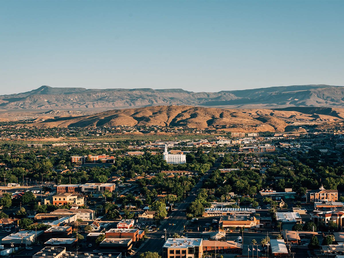 Elevated view of downtown St. George, Utah, with the white LDS Temple visible at center, surrounded by tree canopy, commercial buildings, and a flat-topped mesa in the background.