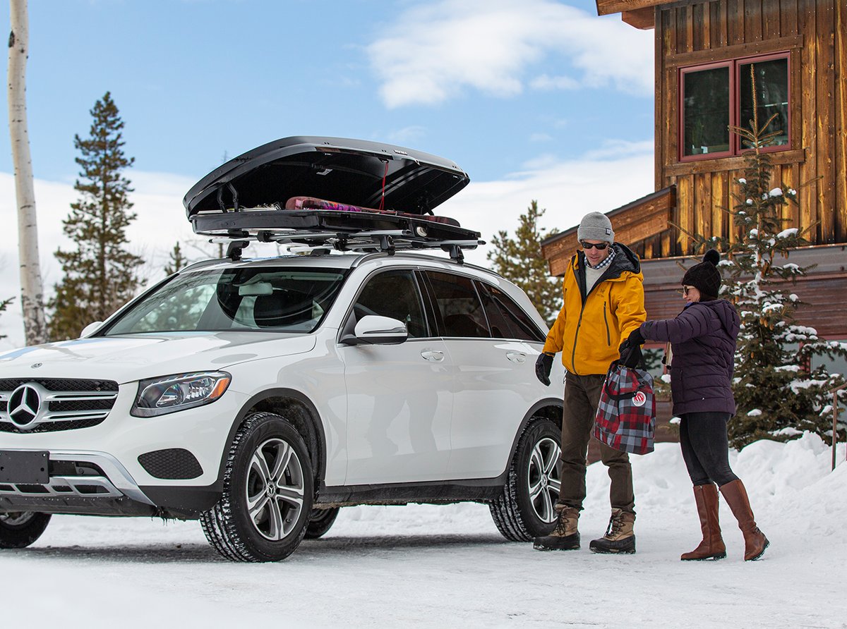 Couple unloading gear from Yakima GrandTour rooftop cargo box on SUV outside snowy mountain cabin
