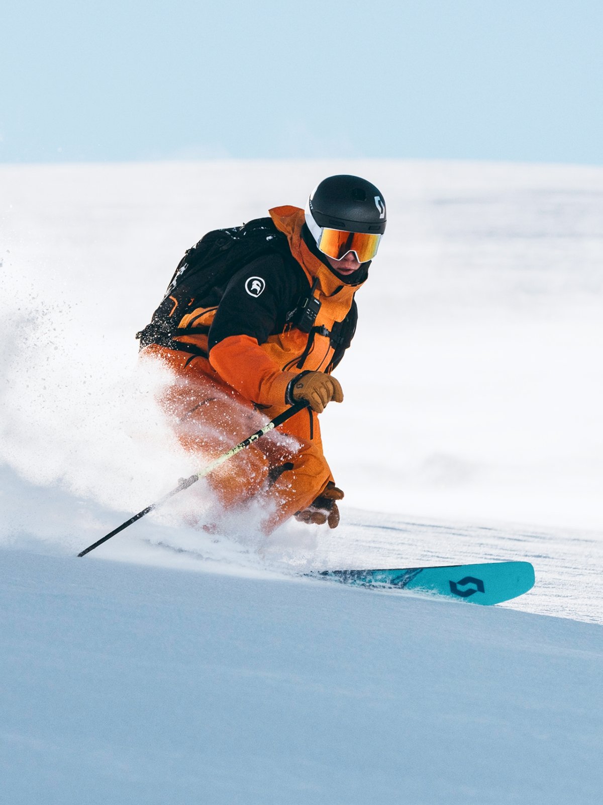 A skier wearing an orange jacket and pants, black helmet, and backpack plows through pow on a bluebird day.