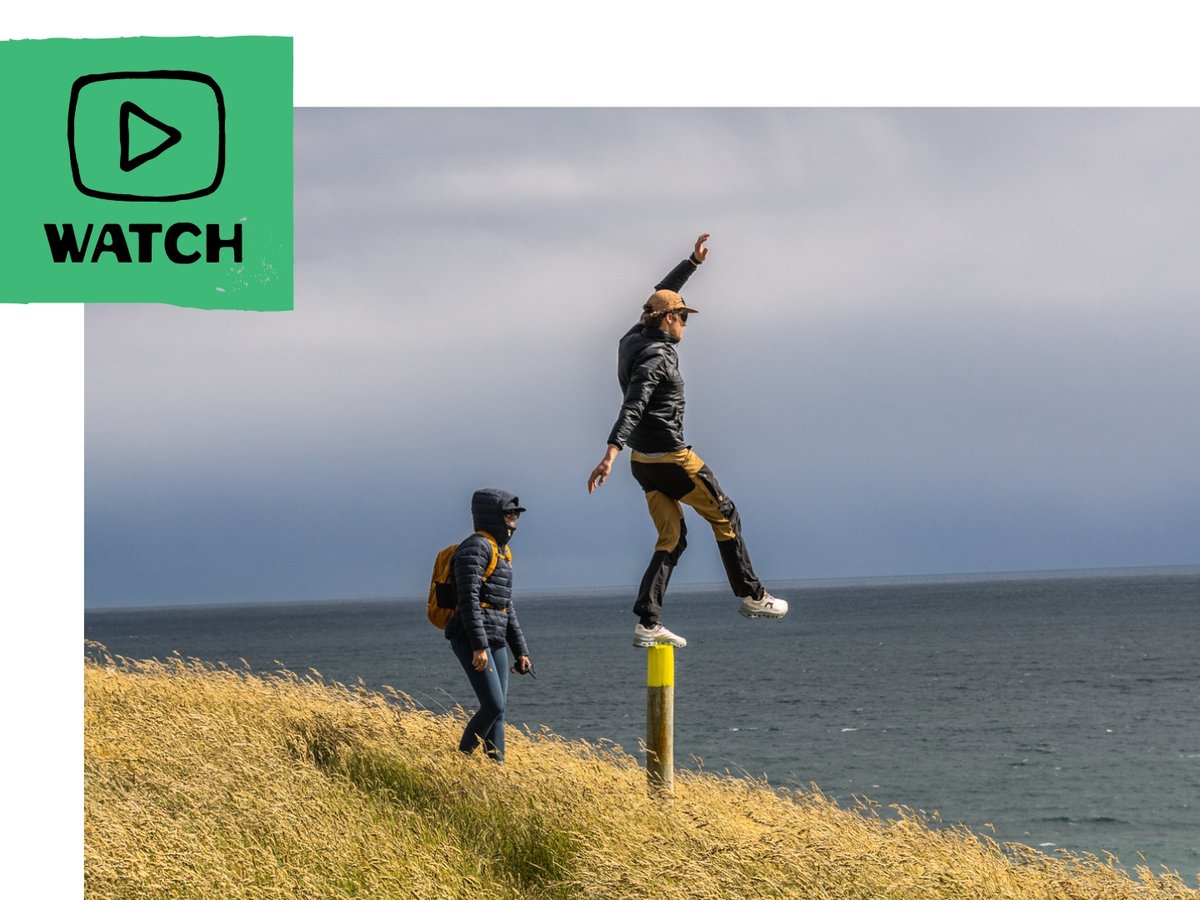 A person in a black jacket and yellow pants jumps off a yellow post near a grassy hillside overlooking the ocean, while another person in a blue jacket and yellow backpack walks nearby. In the top left corner, there  is a green graphic with a play button icon and the word "WATCH."