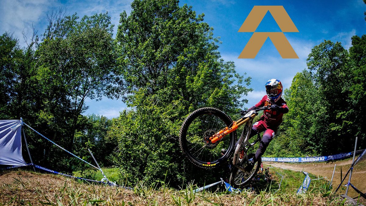 A mountain biker in red gear performs an airborne jump on an orange and black bike during a competitive cycling event surrounded by trees and branded barriers.