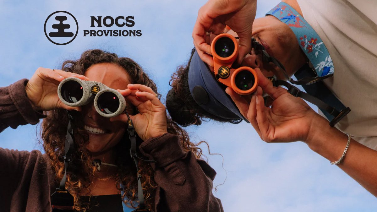 Two people holding binoculars up to their eyes against a blue sky, showcasing outdoor observation gear from NOCS Provisions.
