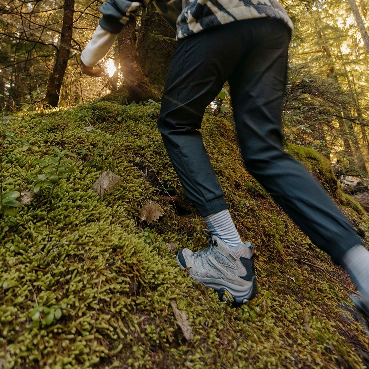 Low-angle close-up of a person's feet in Merrell hiking shoes stepping across a moss-covered log in a forest at golden hour.