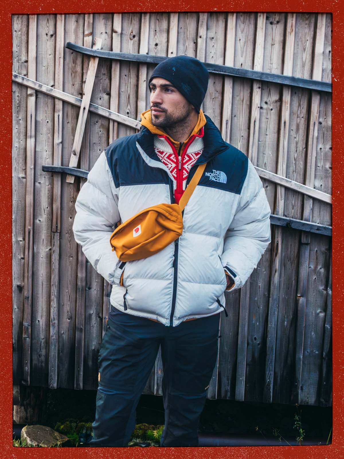 Person in a puffy jacket and beanie with an orange crossbody hip pack, standing against a weathered wooden wall.