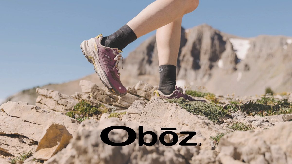 A hiker in burgundy hiking shoes and black socks jumps across rocky terrain with mountains in the background under clear blue sky.