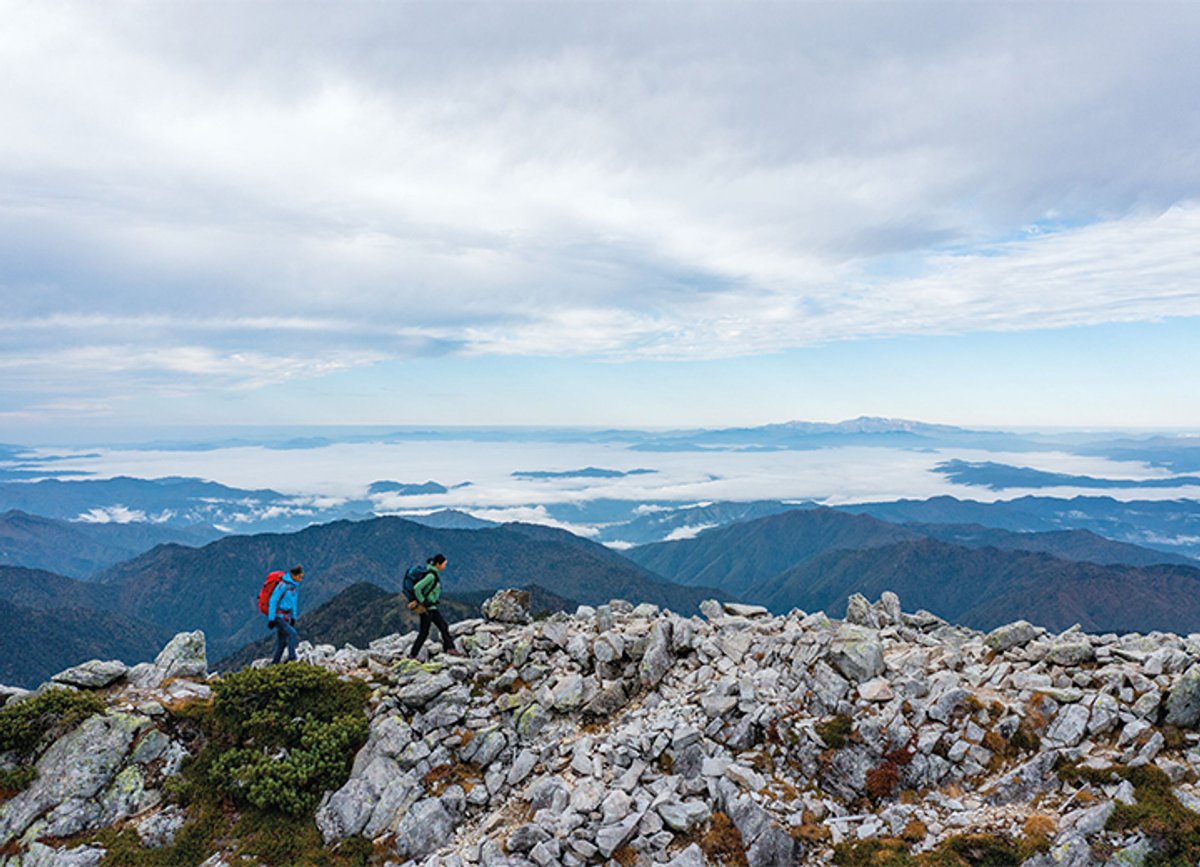 Two hikers walk along a rocky ridge, with a landscape spread out behind them.