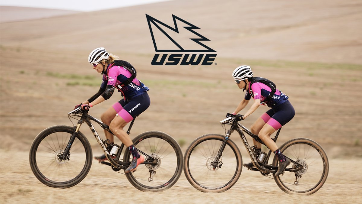 Two female cyclists wearing pink and black USWE gear ride gravel bikes side by side on a dusty desert trail with rolling hills in the background.