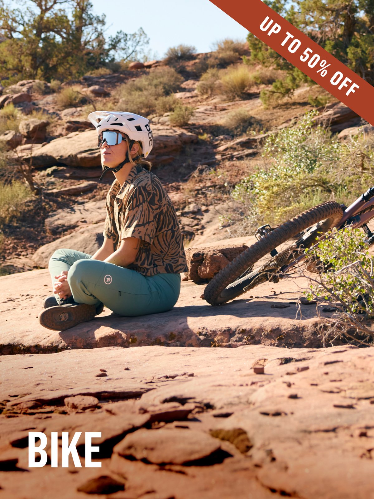 A mountain biker rests cross-legged on red slickrock in a white helmet and teal pants, bike leaned nearby, soaking in the desert quiet. Bike Up To 50% off.