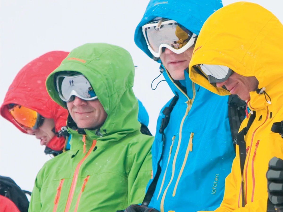 Group of four skiers wearing colorful winter jackets and helmets with ski goggles smiling at the camera in snowy conditions.
