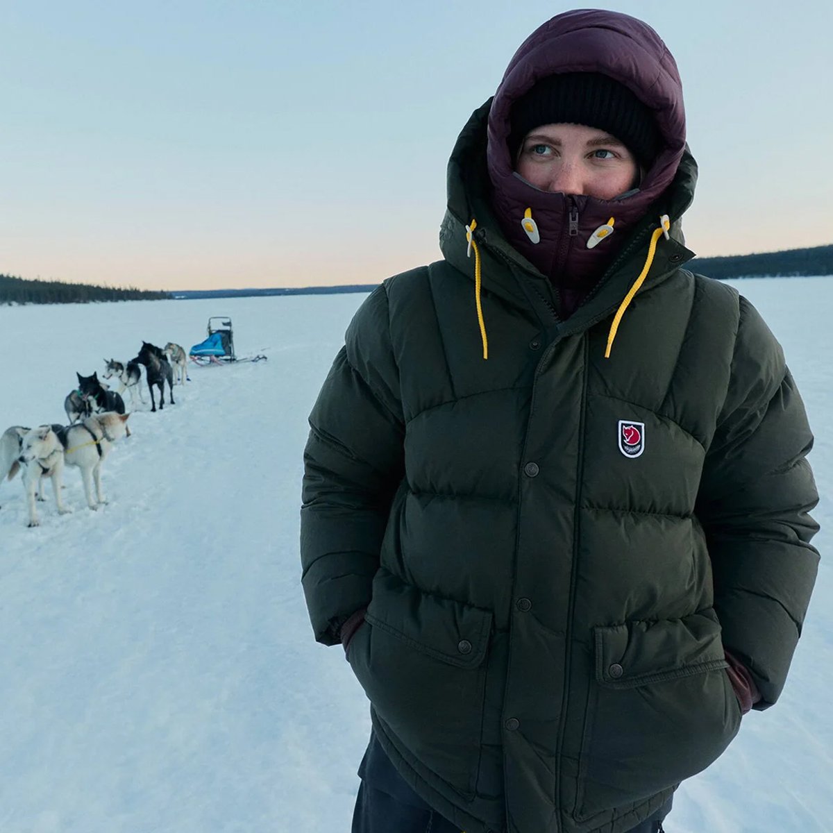 A woman wears a Fjallraven Expedition jacket. She stands on a snowy background with a dog sled behind her.