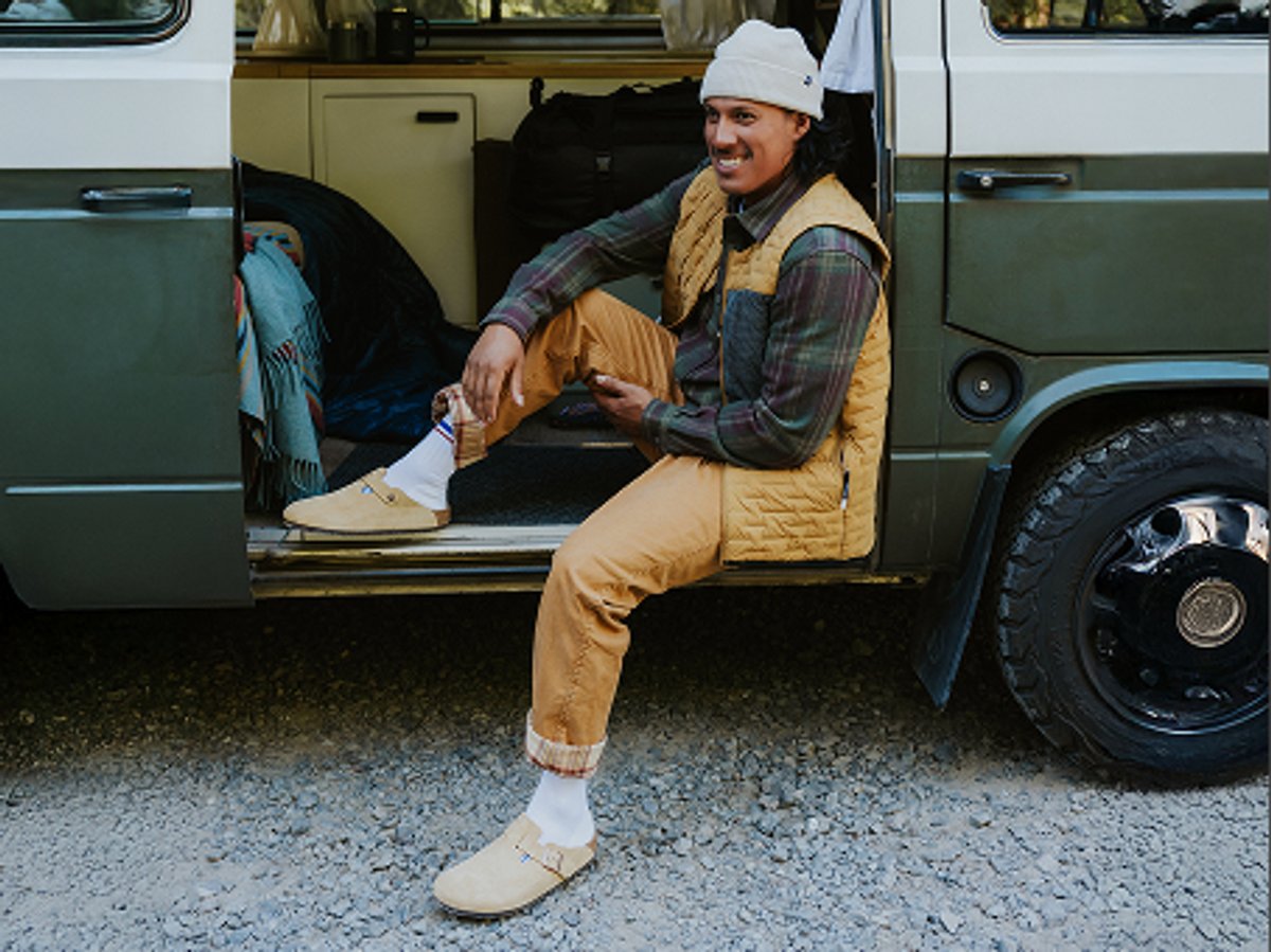 Person sitting in the doorway of a camper van, wearing tan pants, a flannel shirt, and a vest, smiling in an outdoor campsite scene.