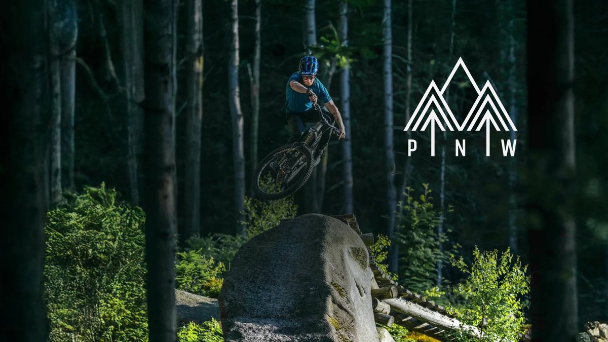 Mountain biker in blue helmet performing an airborne jump off a large rock in a dense forest trail with the PNW logo displayed.