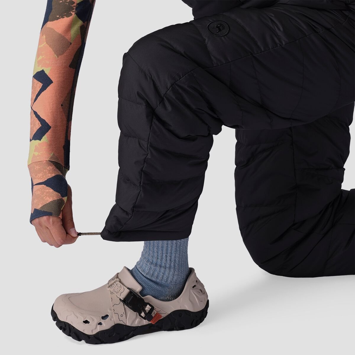 Close-up of a person kneeling to adjust the ankle cuff drawcord on black Stansbury ALLIED Down pants while putting on a shoe.
