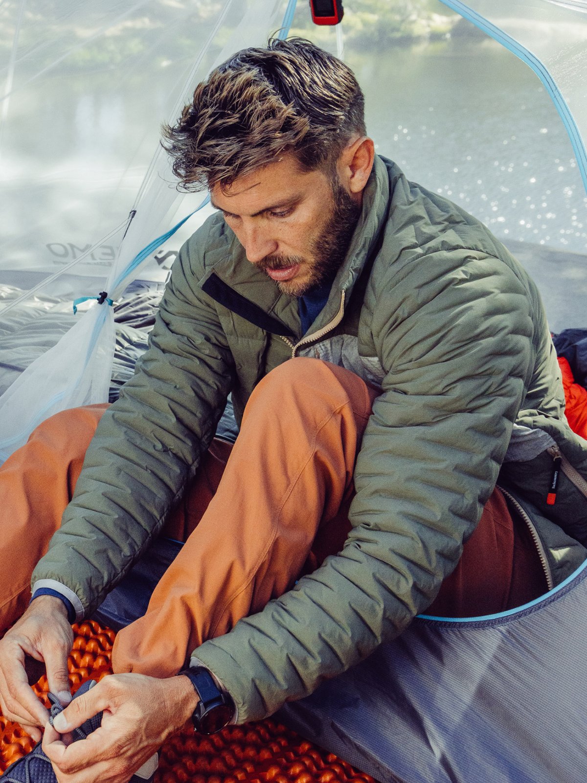 A person in an olive green quilted jacket and orange pants sitting in front of a tent, tying his shoes.