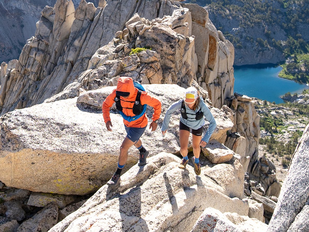Two alpinists wearing tech hoodies, shorts, hiking boots, and running vests scale a bouldery ridge.