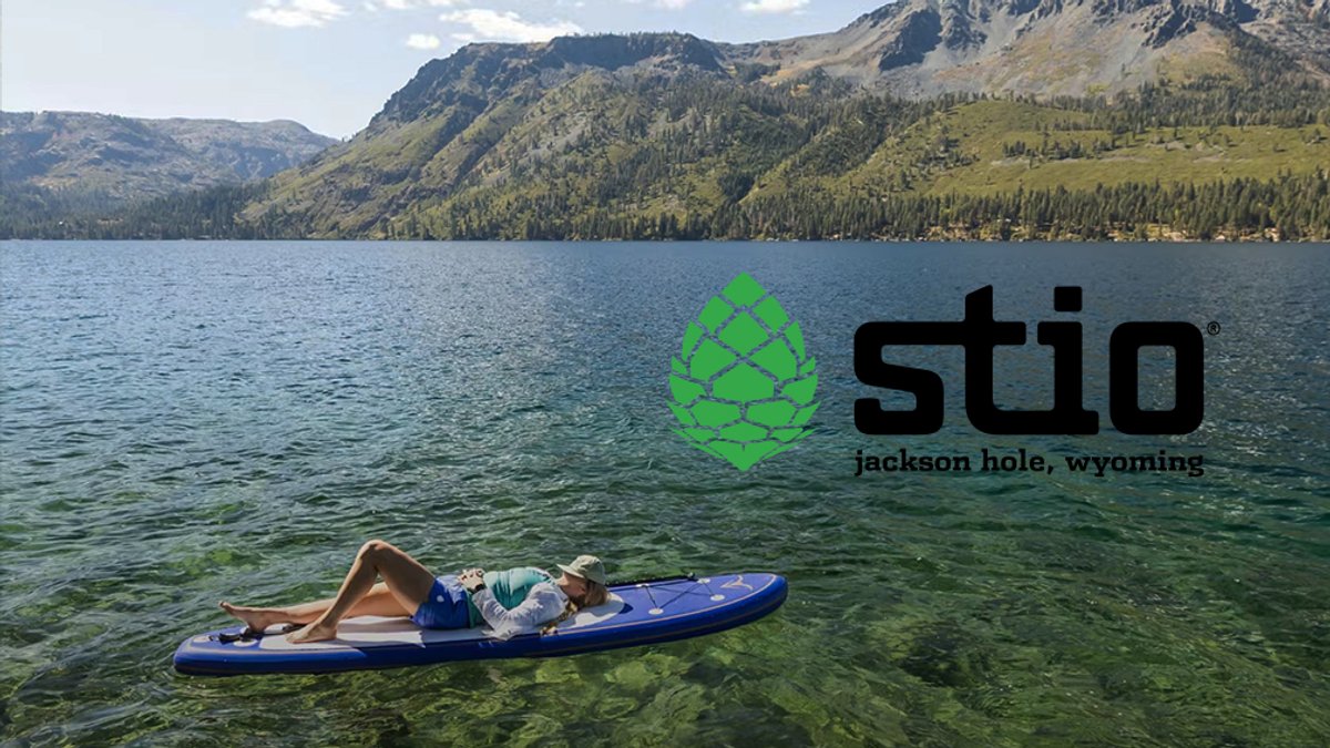 A woman relaxes on a blue paddleboard in crystal-clear alpine lake waters with forested mountains in the background.