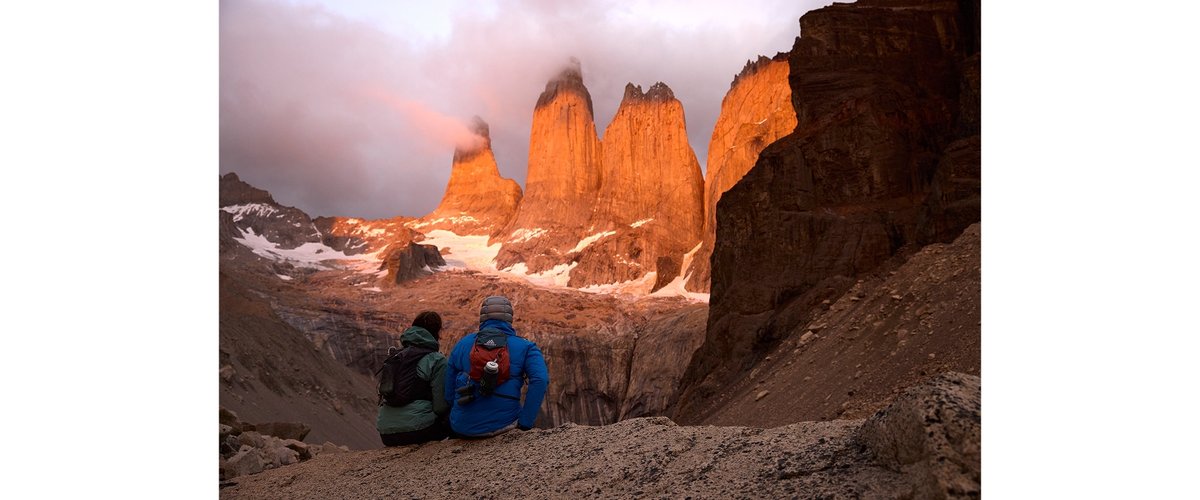 Two people sit on a rocky ledge appreciate the view of the famous Patagonia mountain scape.