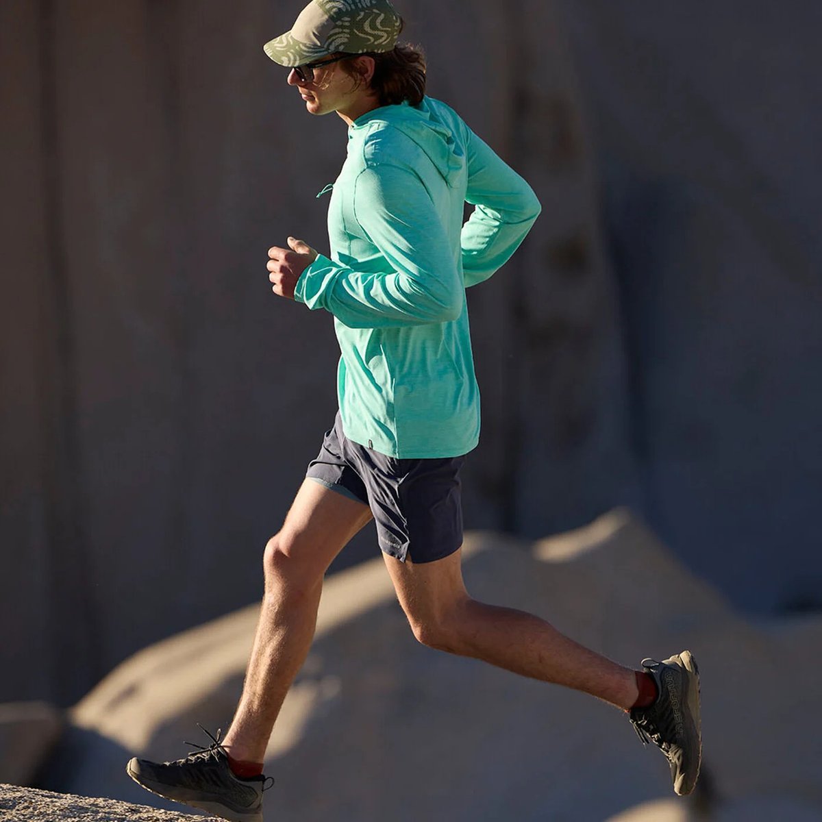 Man jogging outdoors in turquoise long-sleeve running shirt, dark shorts, cap and sunglasses with professional athletic footwear on a sunny day.