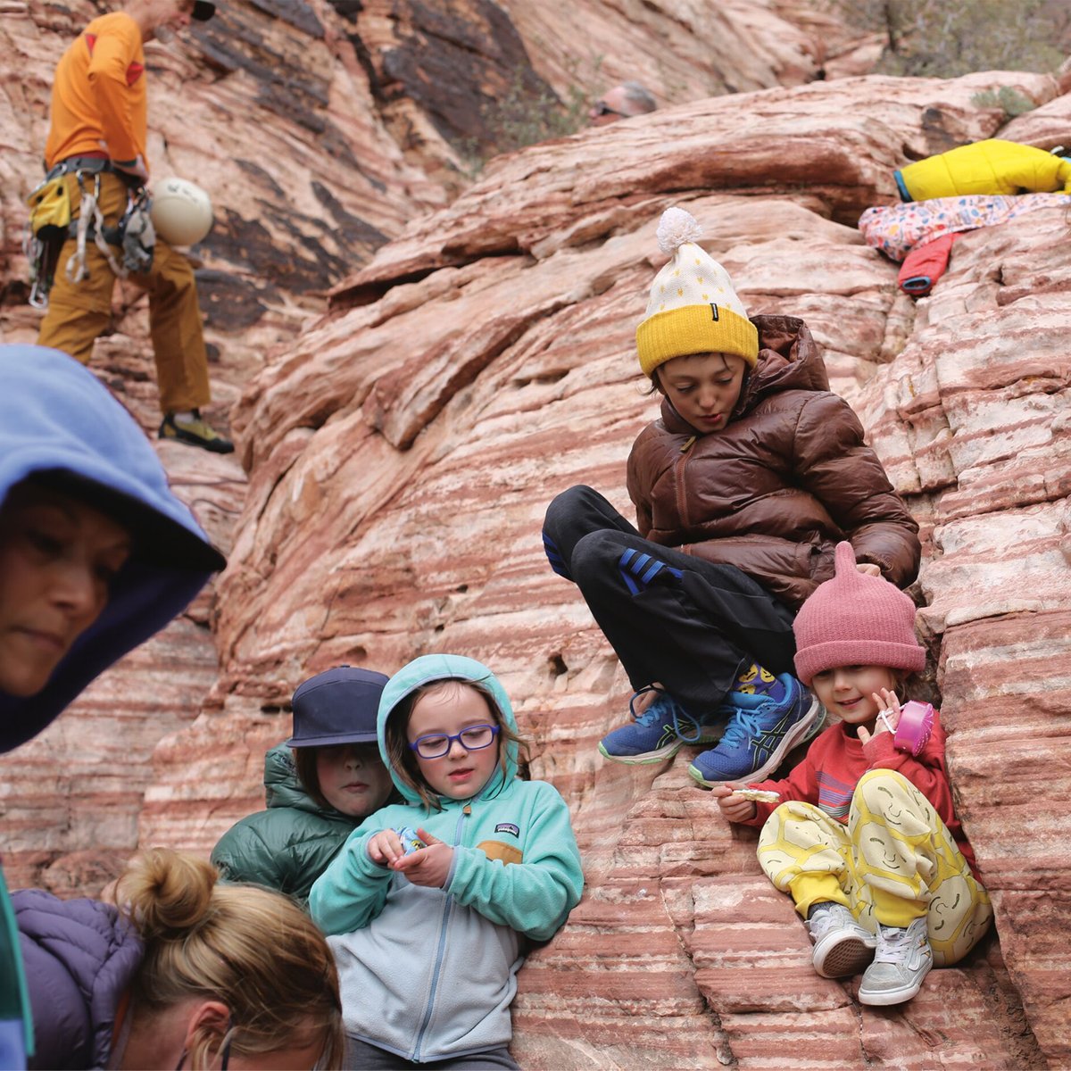 Children and families enjoying rock climbing and outdoor adventures on a natural red rock formation while wearing colorful winter hats and climbing gear.