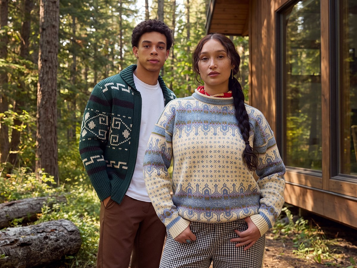 Two people wearing patterned knit sweaters outside a wood cabin surrounded by evergreen forest.