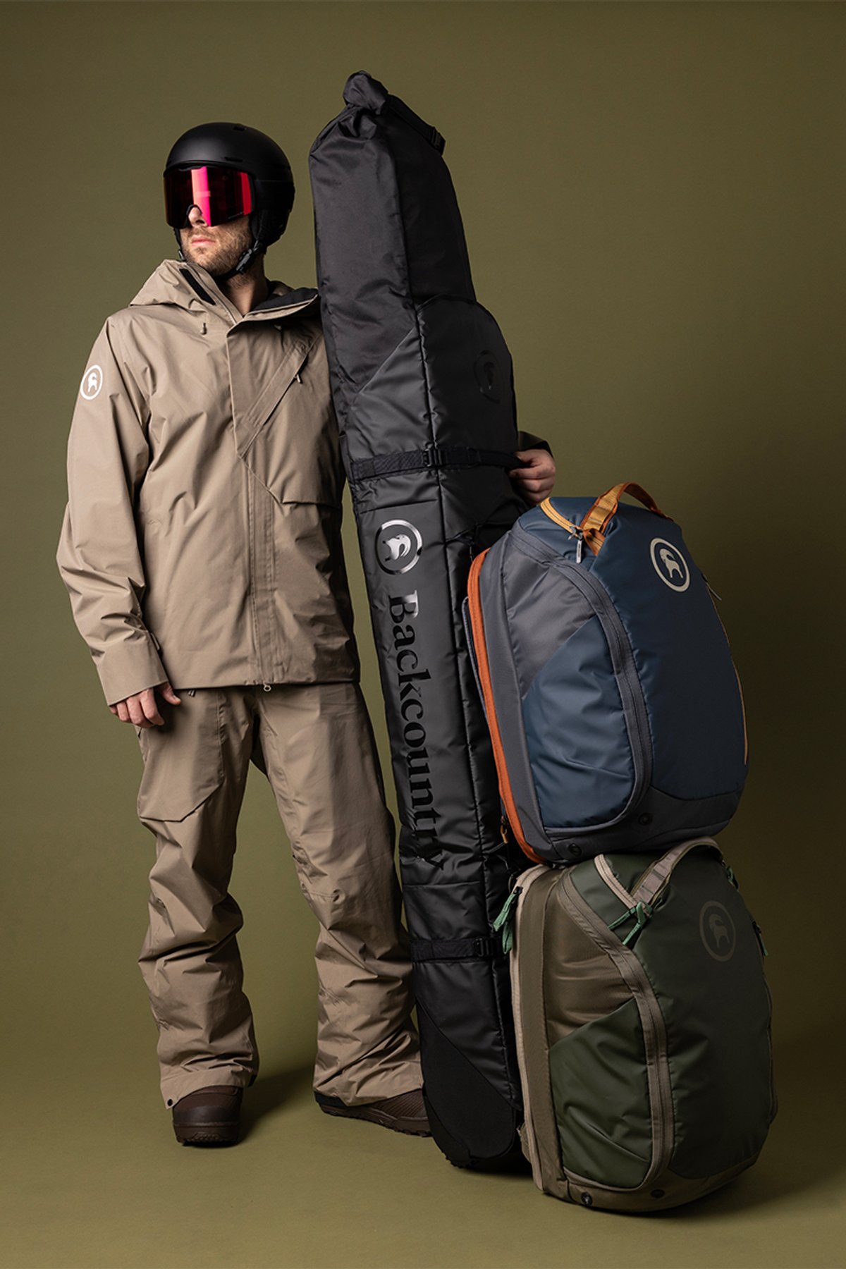 Person in snowabord outerwear wearing helmet and goggles holding rolling ski bag with two boot bags attached.