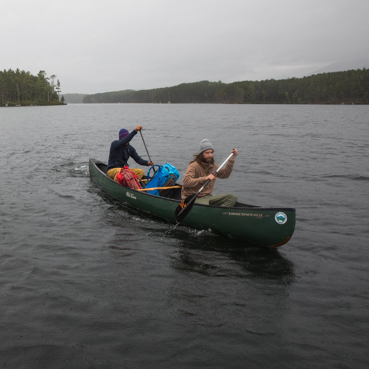 Two paddlers navigate a green canoe across a calm lake surrounded by dense forest in overcast conditions while carrying camping gear and backpacks.