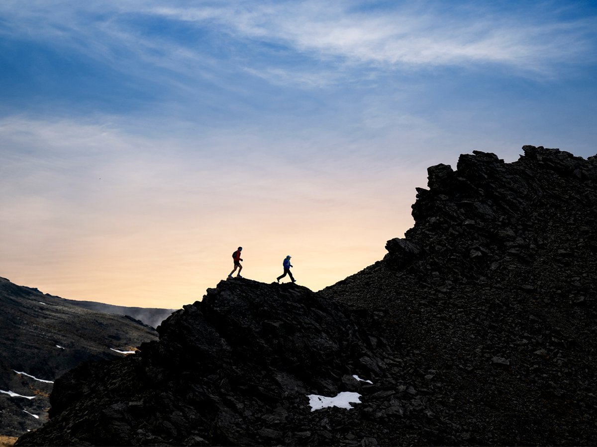 Two hikes walk across a rocky ridge, silhouetted against a sunset.