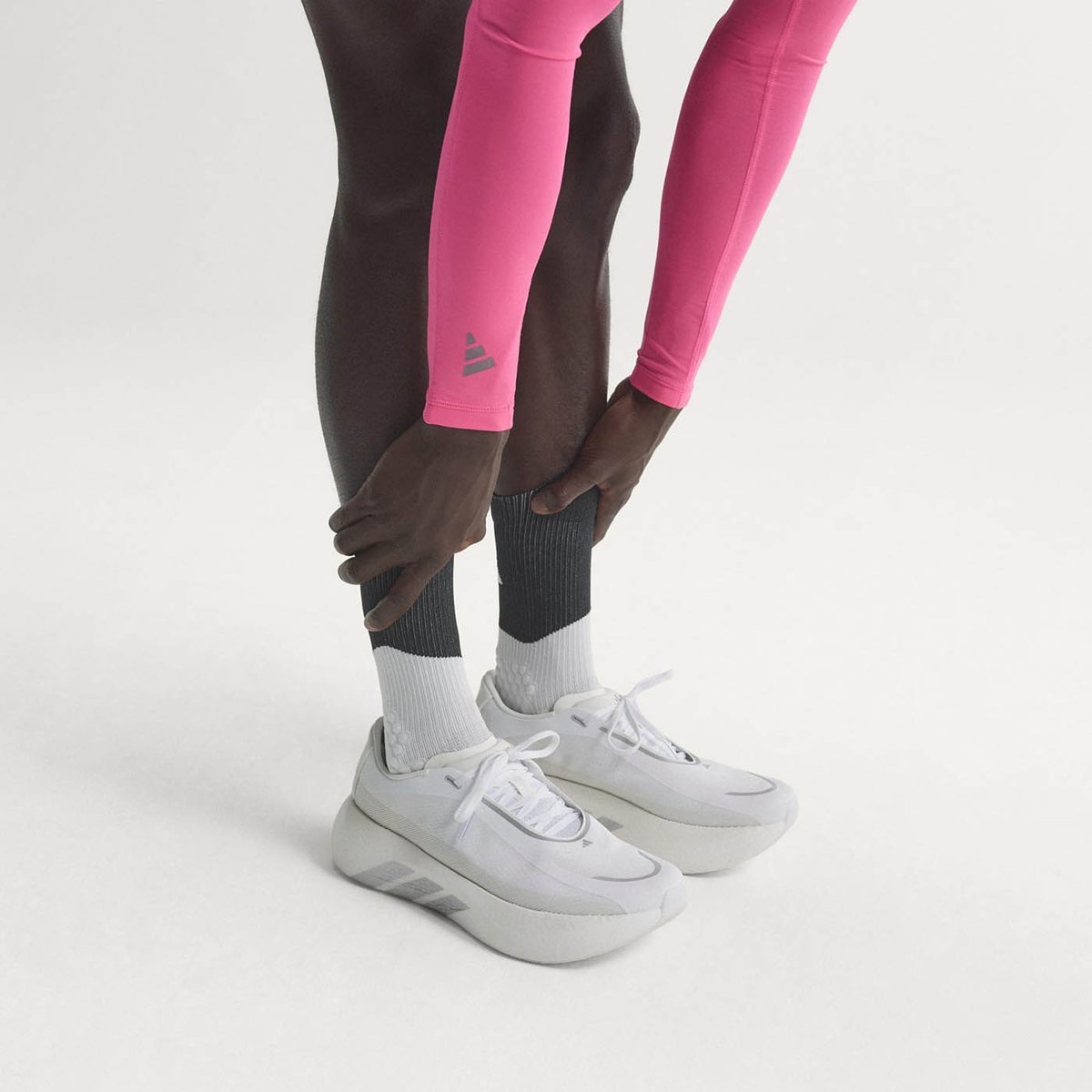 Lower legs of person standing bending over wearing pink Adidas arm sleeves, in white/gray Adidas running shoes and two-tone gray-and-white socks on a white background.